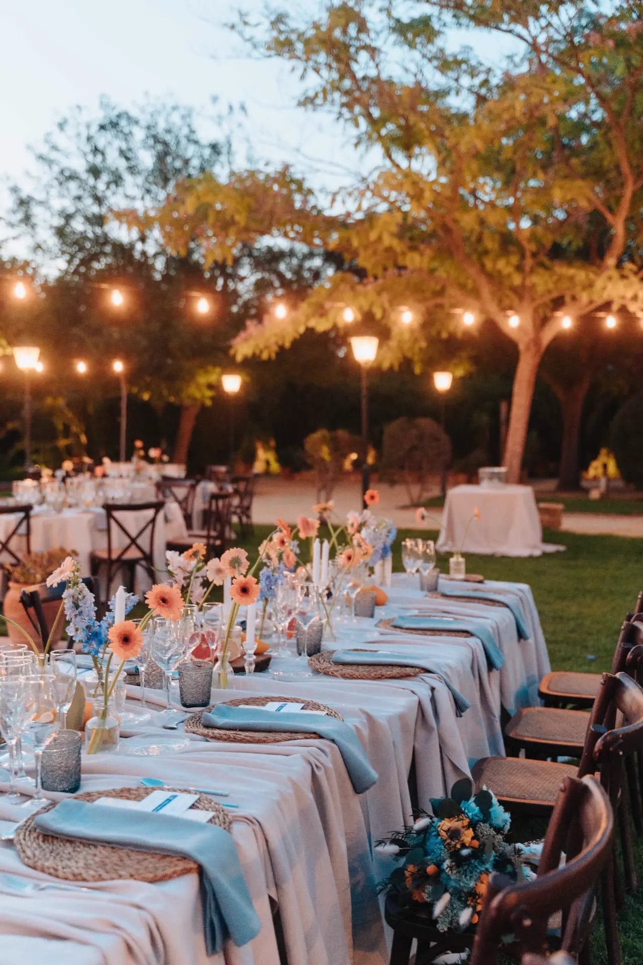 Mesa larga con velas, gerberas coral y servilletas azules bajo las guirnaldas de luces