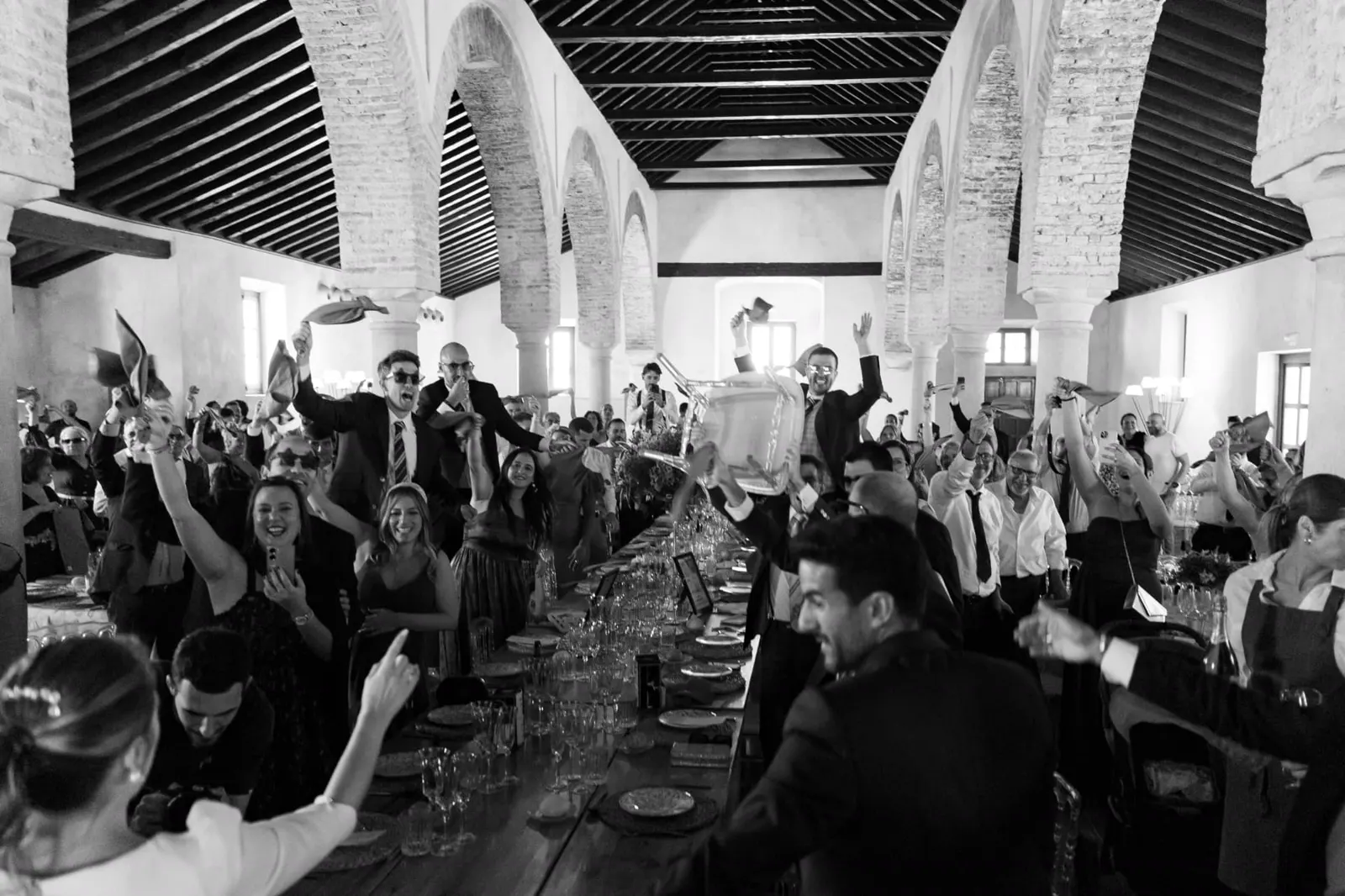 Invitados celebrando con las servilletas en alto en el salón abovedado, en blanco y negro