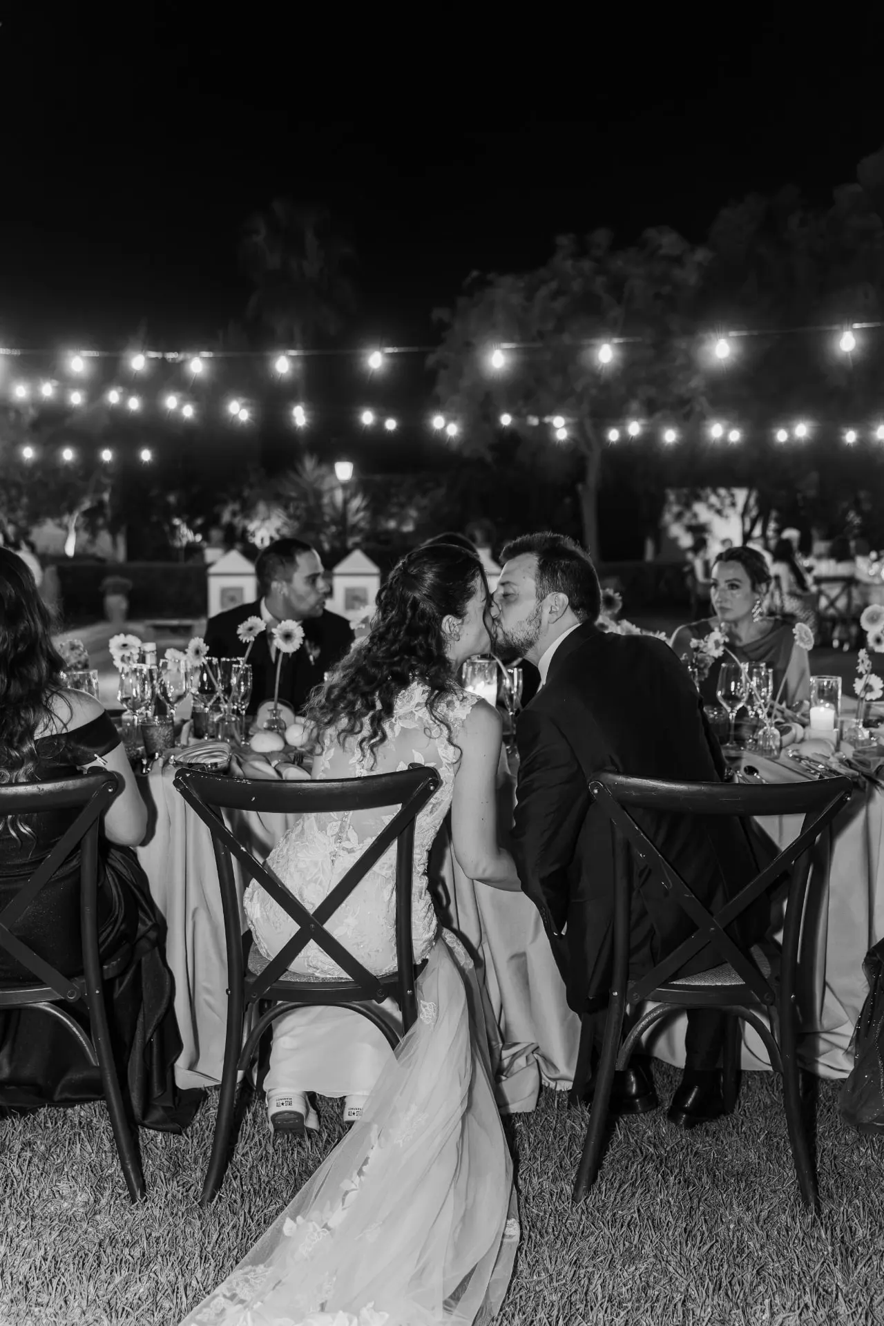 Ana y Manuel besándose en la mesa presidencial bajo las guirnaldas de luces, en blanco y negro